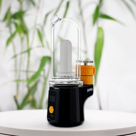 Das Bomb Sykloud E Rig (farbige Option: schwarz) mit einem Glasbecher und einer orangefarbenen Beschichtung der Heizkammer, abgebildet auf einer weißen Oberfläche vor einem unscharfen Hintergrund aus grünen Blättern.